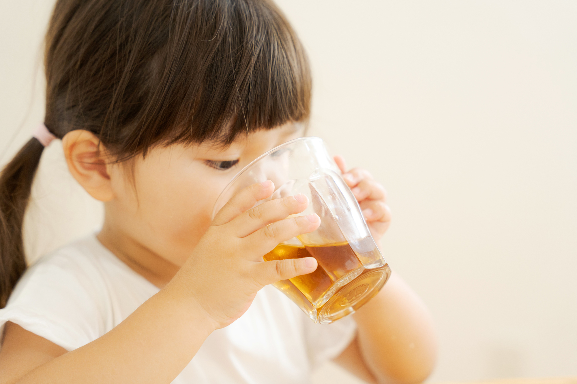透明なコップを持ち、お茶(または水)をごくごくと飲んでいる幼い女の子のアップ写真。日常的な水分補給でむし歯を防ぐライフスタイルを表現したイメージ画像。