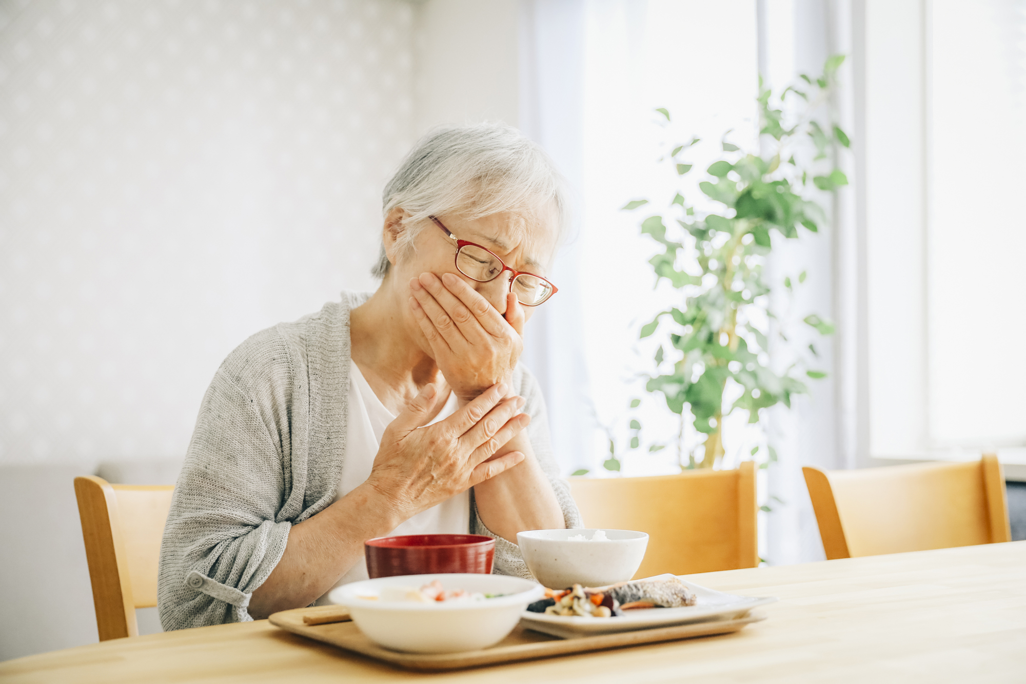 食卓で食事中に手で口元と胸元を押さえ、むせたり飲み込みにくそうに苦しそうな表情を浮かべている高齢女性の様子。