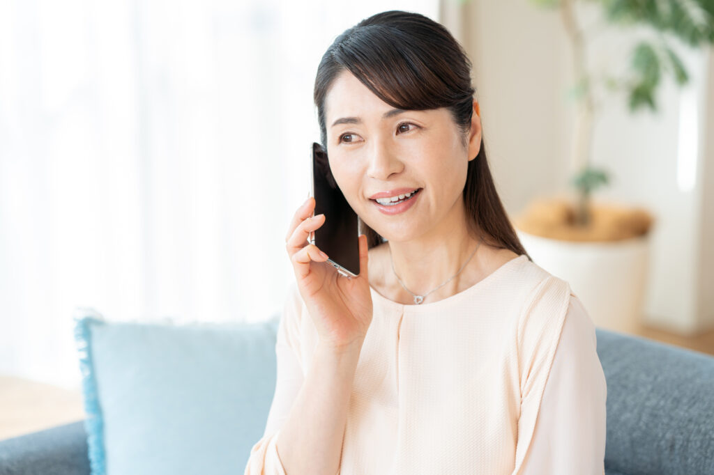 明るいリビングのソファに座り、スマートフォンを耳に当てて笑顔で電話問い合わせをしている中高年女性のイメージ写真。