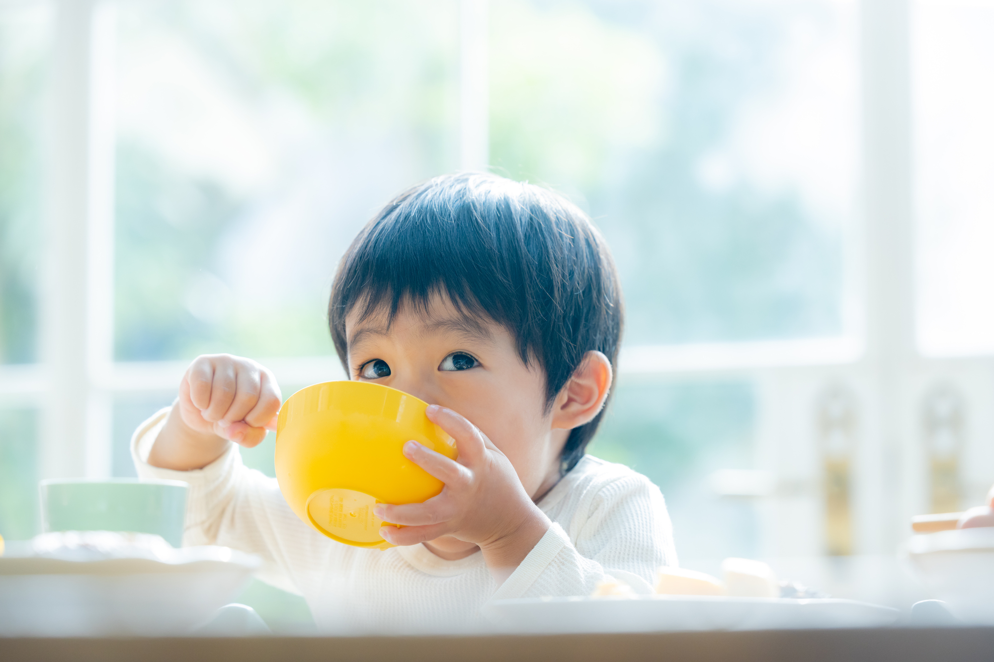 器を両手で持ち、食事やおやつを食べている小さな男の子の様子。ダラダラ食べを防ぎ規則正しい食生活を送る大切さを表現したイメージ画像。