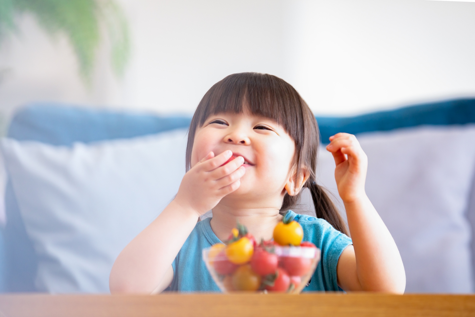 器に盛られたカラフルなミニトマトを嬉しそうに食べている幼い女の子の笑顔の写真。むし歯になりにくくお口に残りにくいおやつの選び方を説明するイメージ画像。
